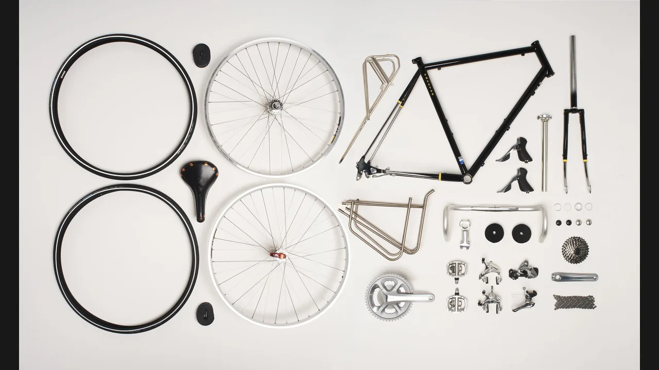 A bicycle frame, wheels, saddle, handlebars, crankset and other parts laid out flat on a white background