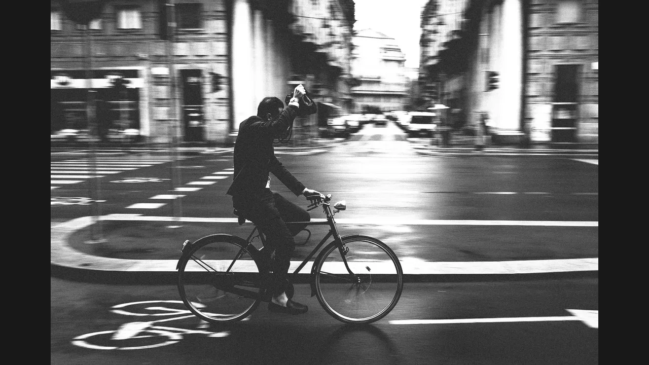 A man holding a camera up to photograph while riding a bicycle on a city street